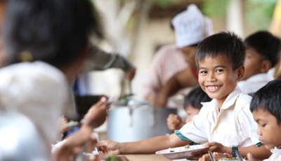 Boy smiles at the camera, surrounded by his schoolmates eating their free school meal in Cambodia