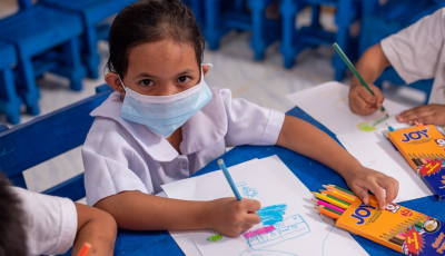 Girls from the Philippines wearing a face mask and school uniform looks up to the camera while drawing at school