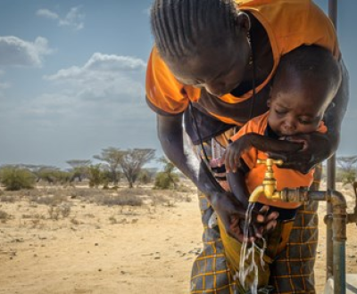 Mother from Uganda helping her young son drink water from an outdoor water tap