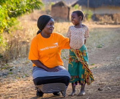 World Vision community development worker in an orange branded tshirt crouches down and smiles and a young girl who is smiling back at her in Malawi