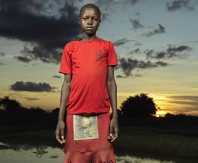 Girl form South Sudan wearing a bright red dress pictured standing alone at sunset in a displacement camp where she lives