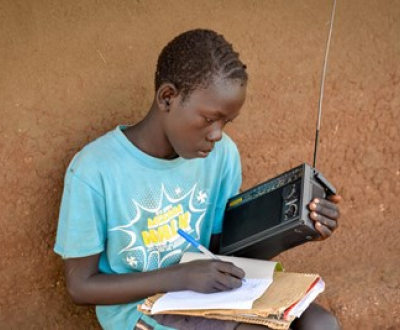 Child from Uganda sits and writes on paper with one hand, holding a radio in the other