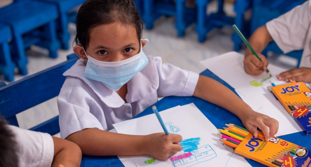 Girls from the Philippines wearing a face mask and school uniform looks up to the camera while drawing at school