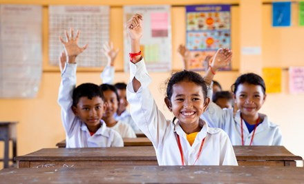 A class of children from Cambodia pictured wearing school uniforms in a classroom and all raising their hand and smiling