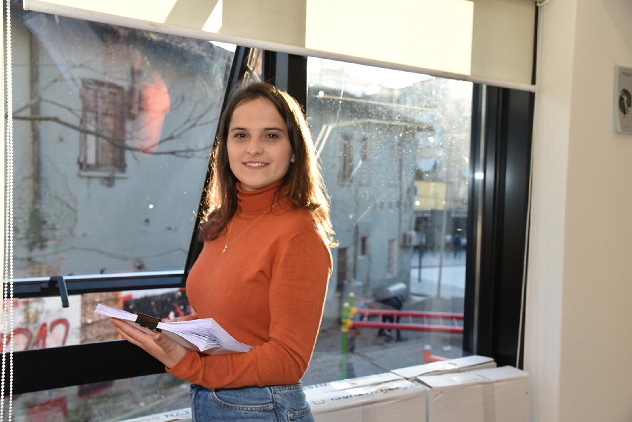 Girl from Albania standing next to a window and holding a book while smiling to the camera