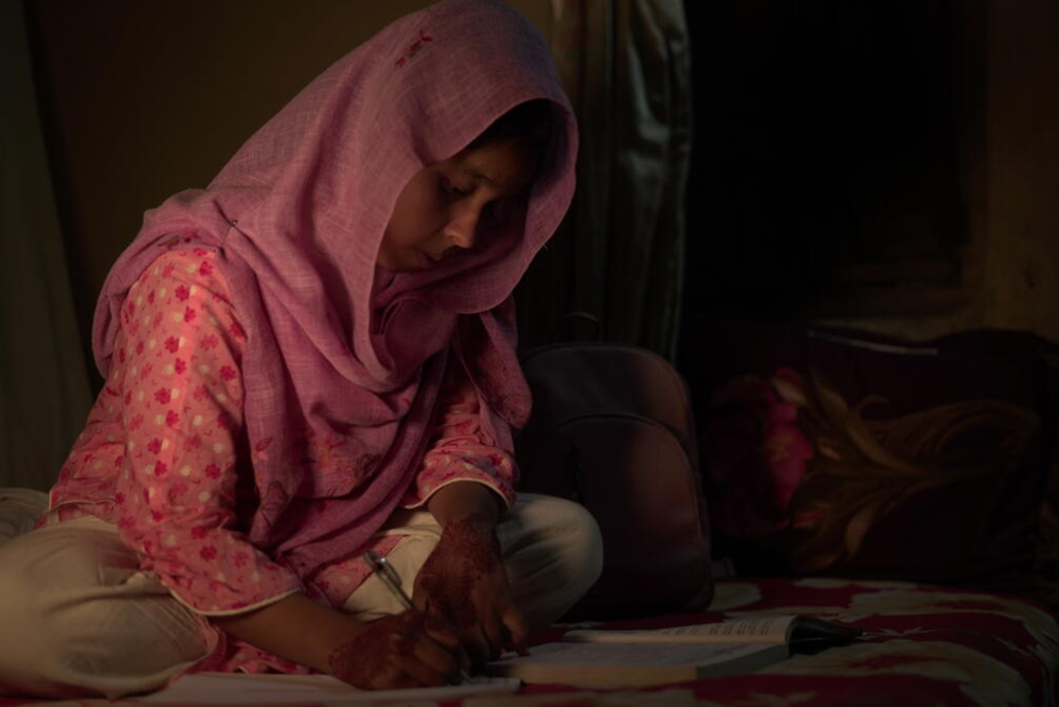 Girl from Bangladesh sitting on the floor in a dark room, writing notes in a notebook