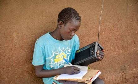 Child from Uganda sits and writes on paper with one hand, holding a radio in the other